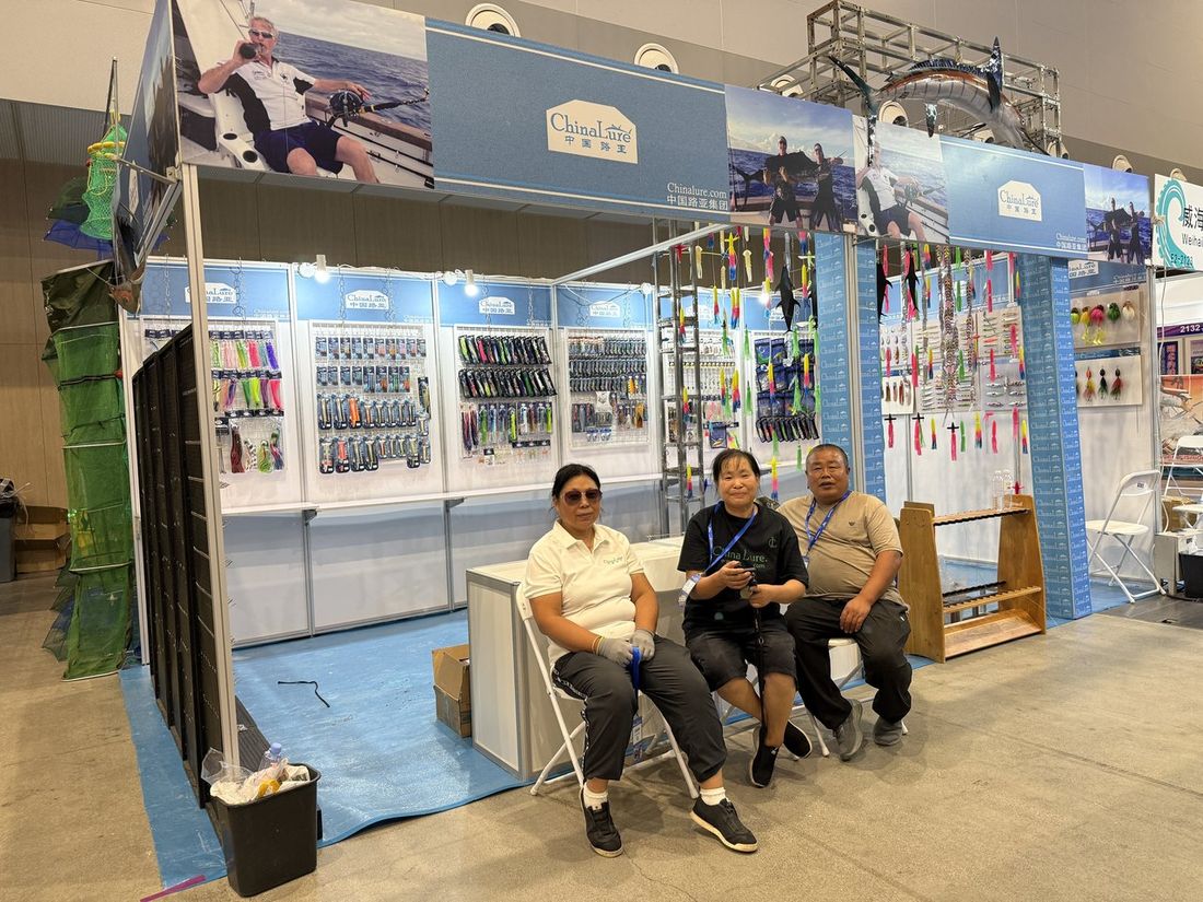 The Chinalure booth at the Weihai fishing tackle expo, with walls of soft lures, squid jigs and trolling teasers and three staff members sitting in front